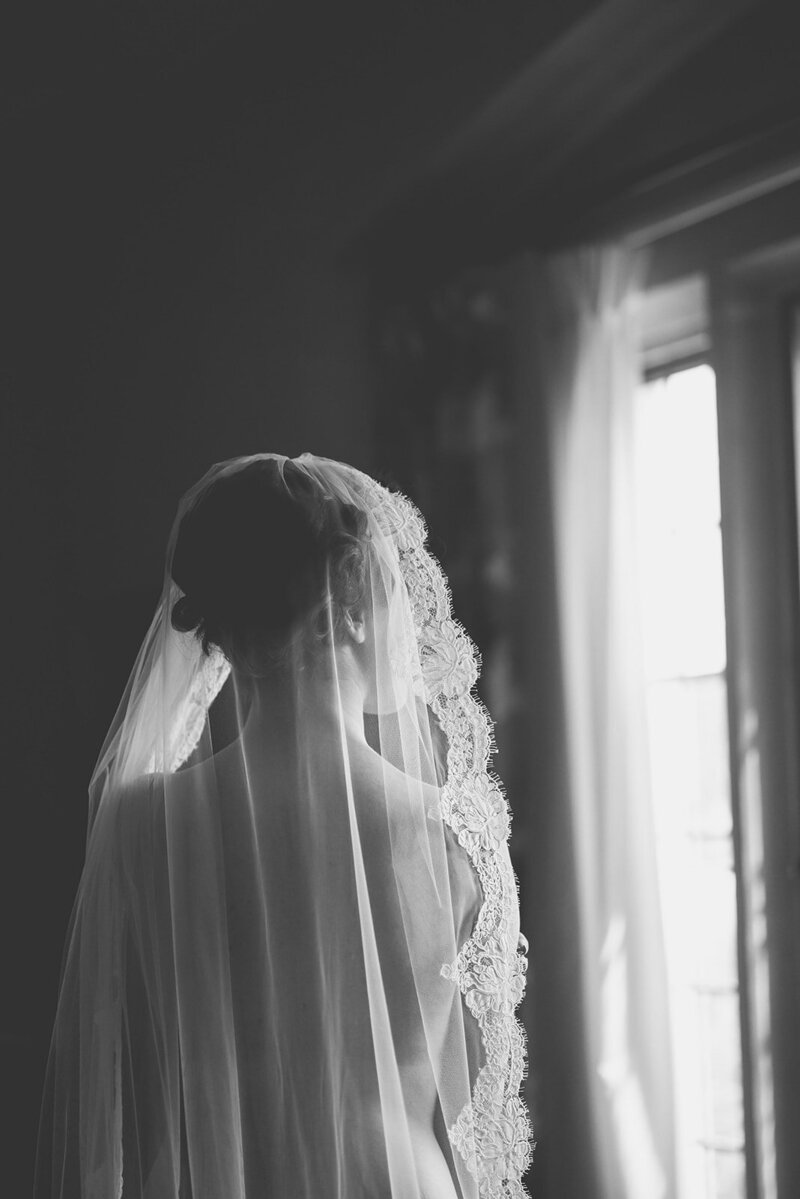 A bride stands in front of a softly lit window, her lace veil catching the light as she takes a quiet moment before the ceremony.