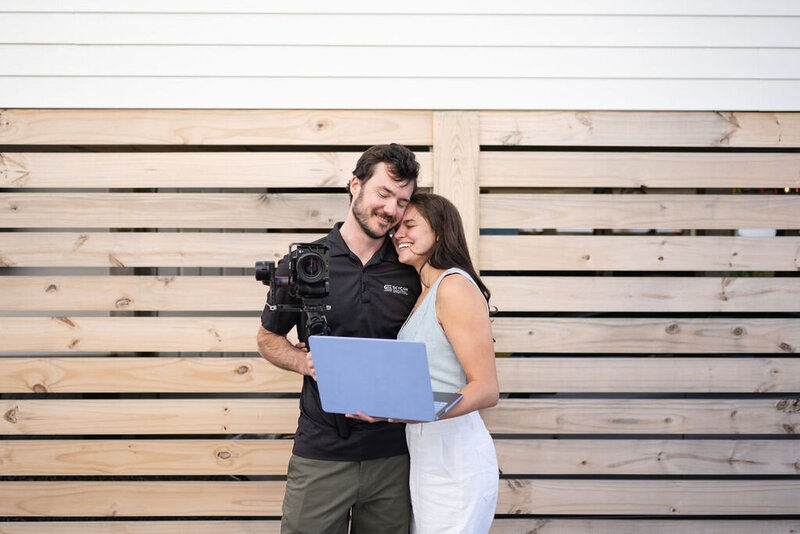 couple stand together holding camera and laptop with wooden slats in the background