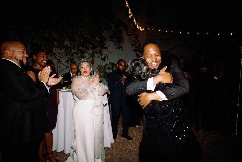 groom hugging wedding guest with his bride smiling and clapping behind him