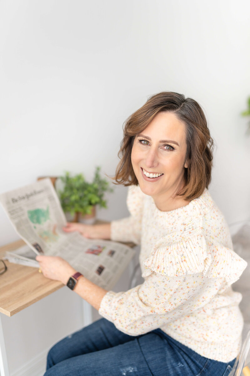Woman smiling from desk and holding newspaper.