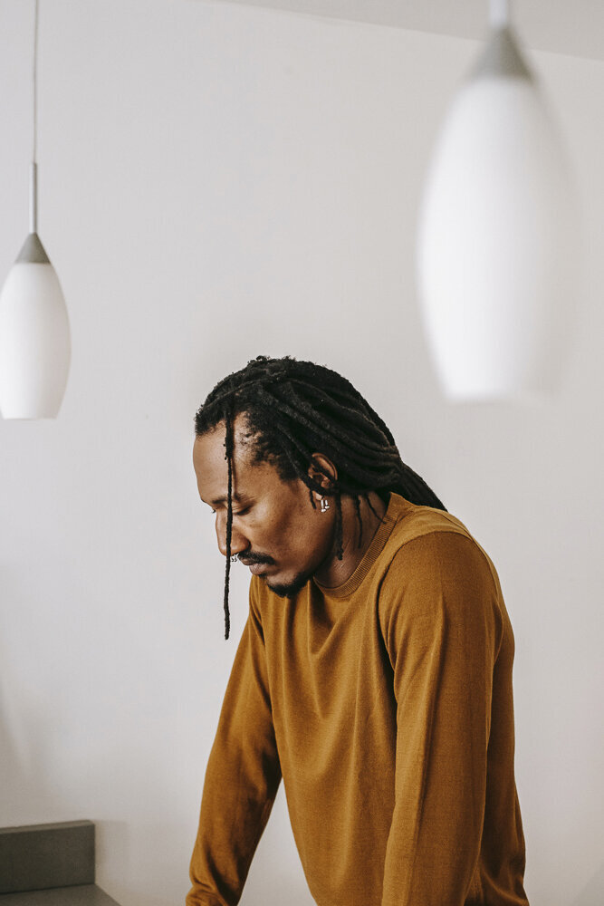 This image shows a black man with dreadlocks pulled back into a low ponytail, wearing a yellow sweater. He looks down toward the counter where his hands are resting, with a tired expression on his face.