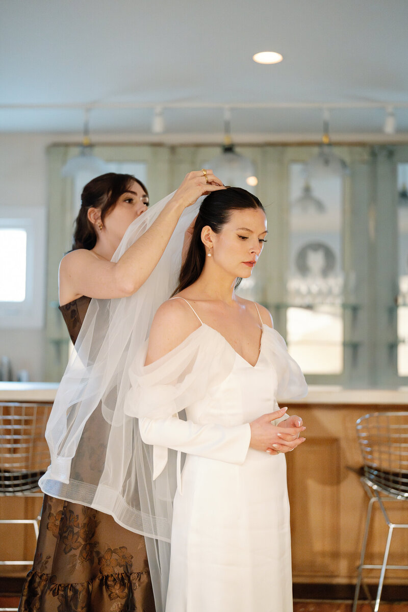 Bridesmaid delicately places veil on bride in serene preparation moment