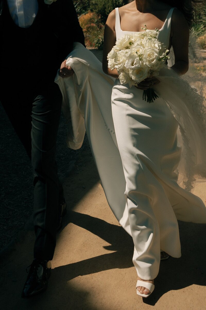 Bride walking holding her bouquet while groom walks beside her holding the train of her dress