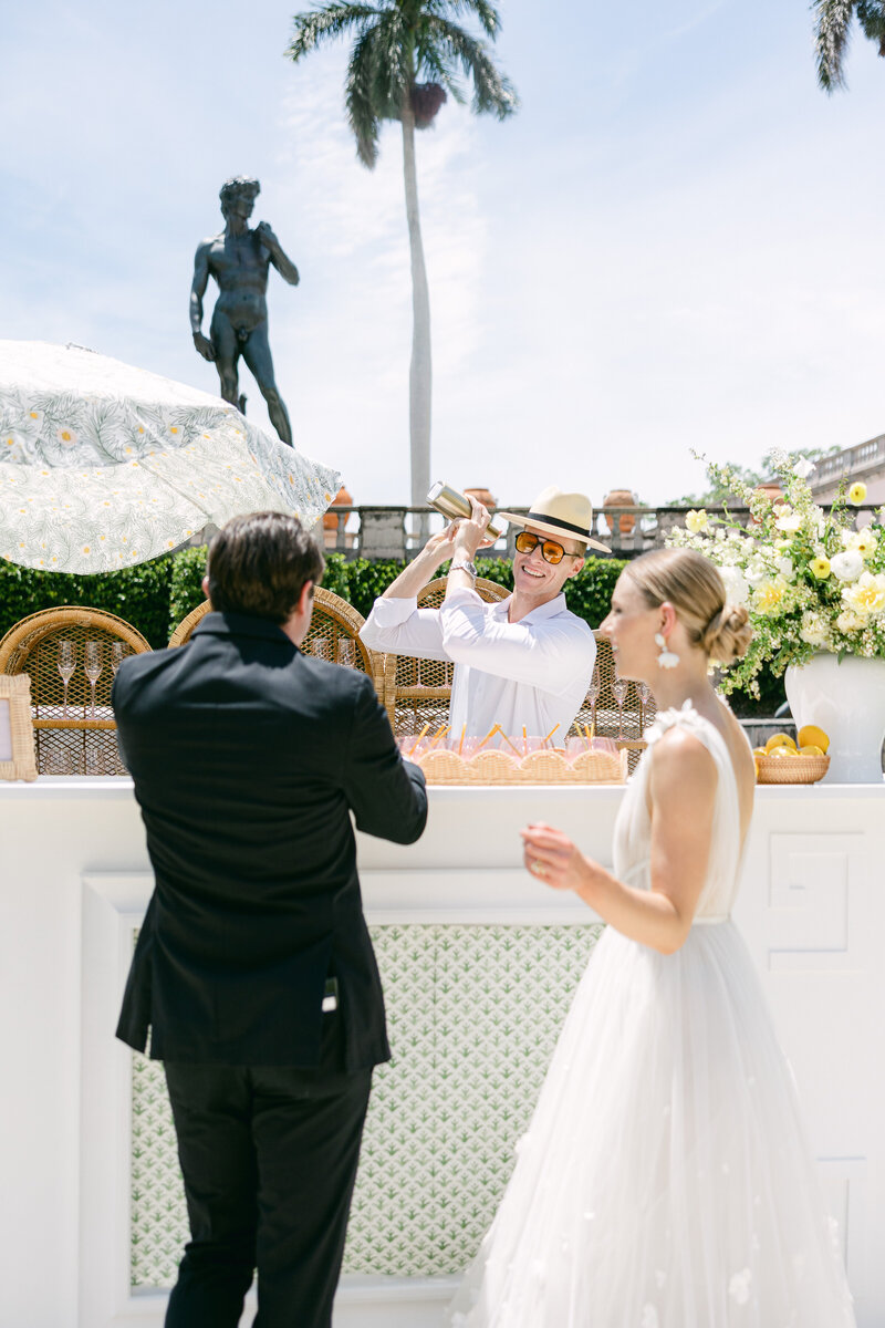 A stylish bride and groom enjoy a lively cocktail hour at their Sarasota Ringling Museum wedding, with a bartender in a Panama hat shaking drinks against a backdrop of elegant décor, lush florals, and classical statues. A luxury wedding captured by Amia Marcell Photography.


