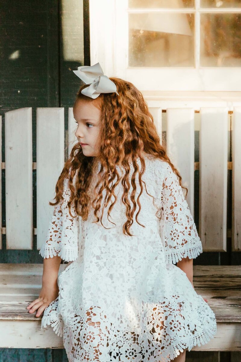 Girl with coffee colored hair in a white dress sitting on a white wooden swing.