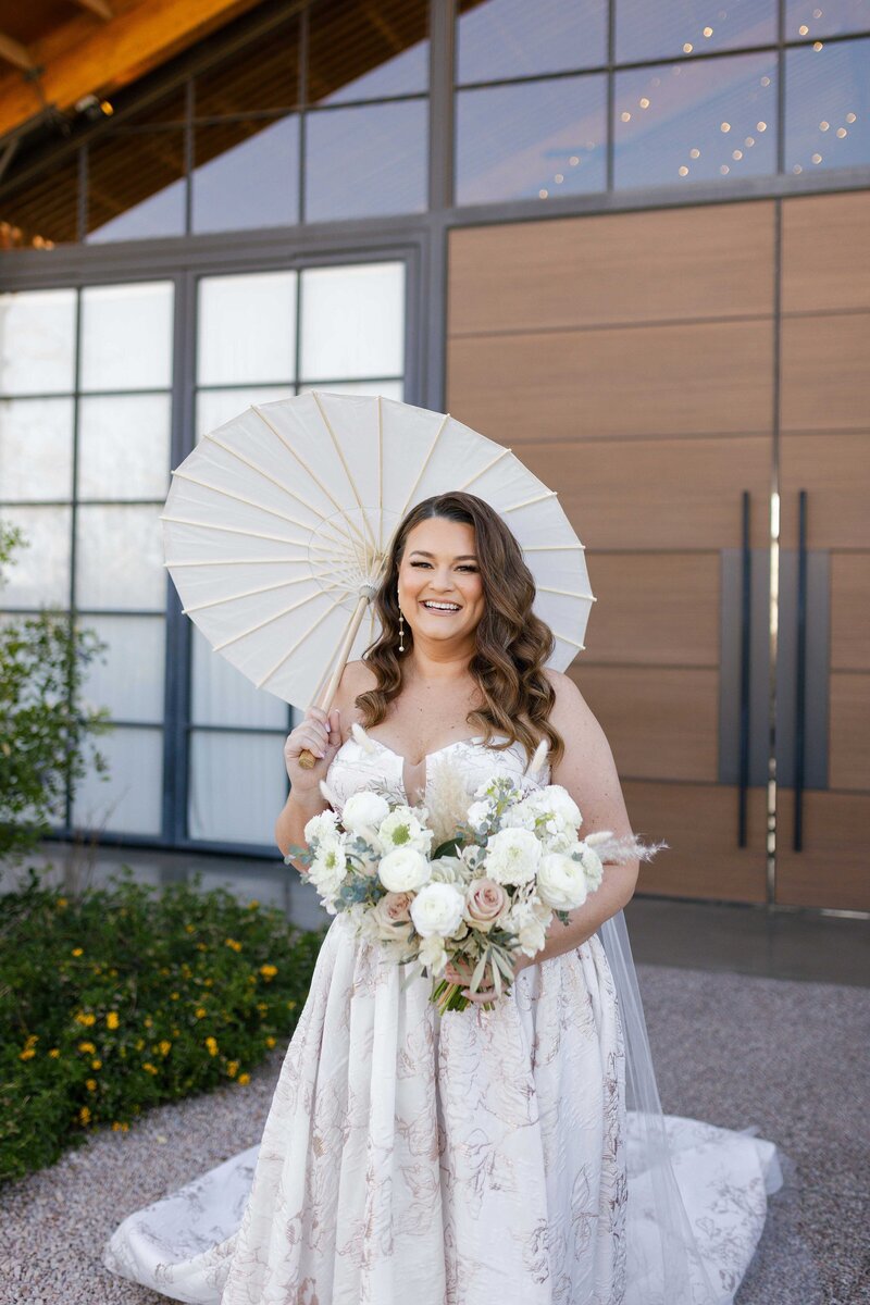 Bride smiles while holding white parasol and bouquet.