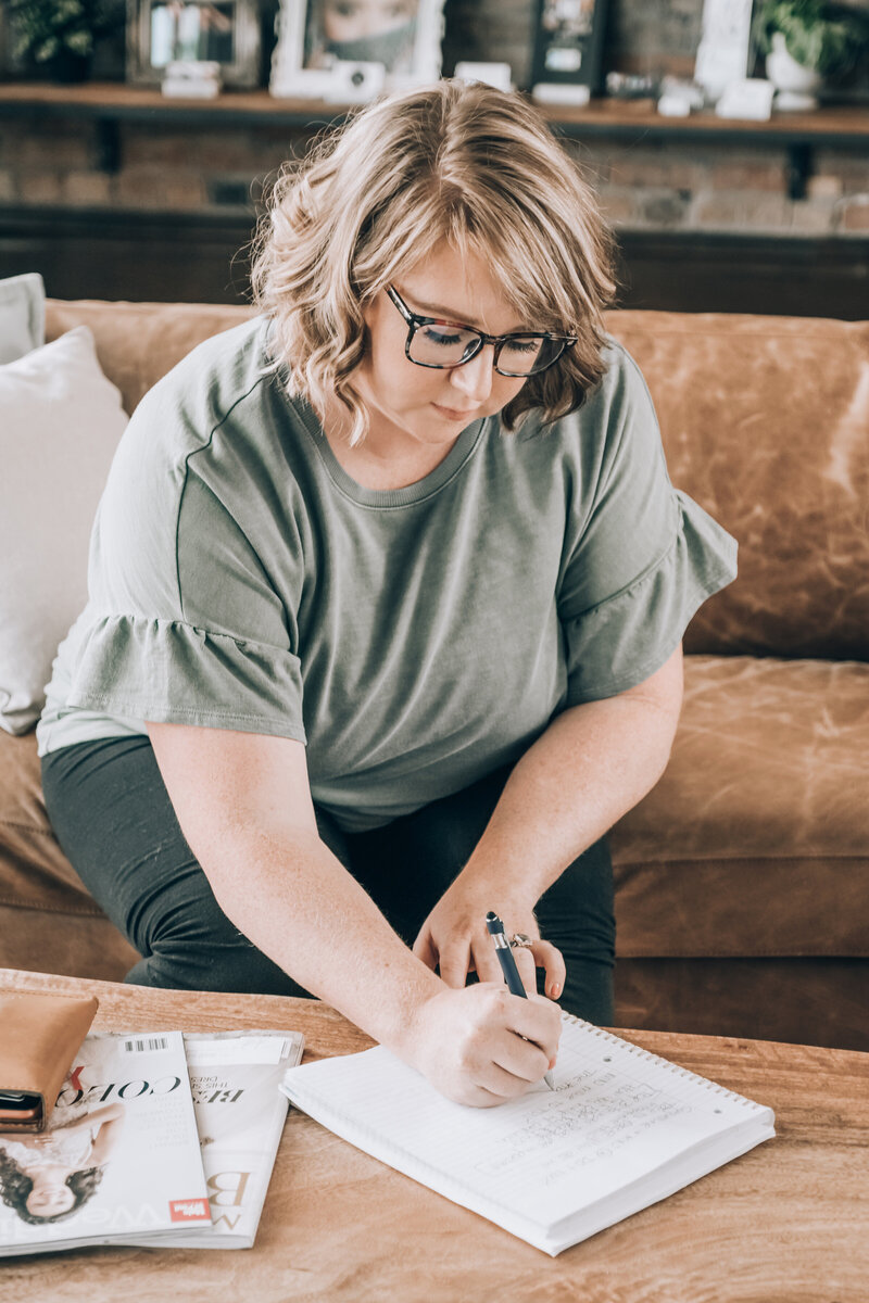 woman in green top sits on a leather couch and works on a notepad on a table
