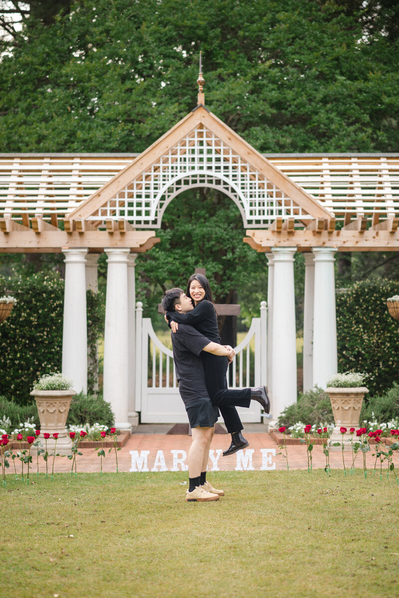 Charlotte proposal photographer captures a joyful engagement moment at Daniel Stowe Botanical Garden, with a man lifting his partner in front of a 'Marry Me' sign, surrounded by red roses.