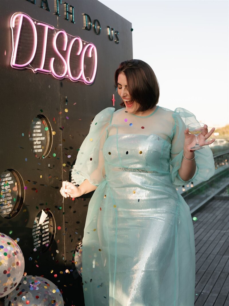 woman happy in metallic teal dress at a wedding on a terrace popping a ballon that is filled with multicolored confetti to find her seat on a black escort wall.