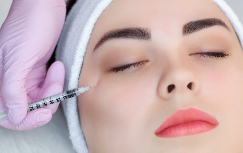 Closeup image of a woman receiving a neurotoxin Botox injection under her right eye from an esthetician wearing a pink glove
