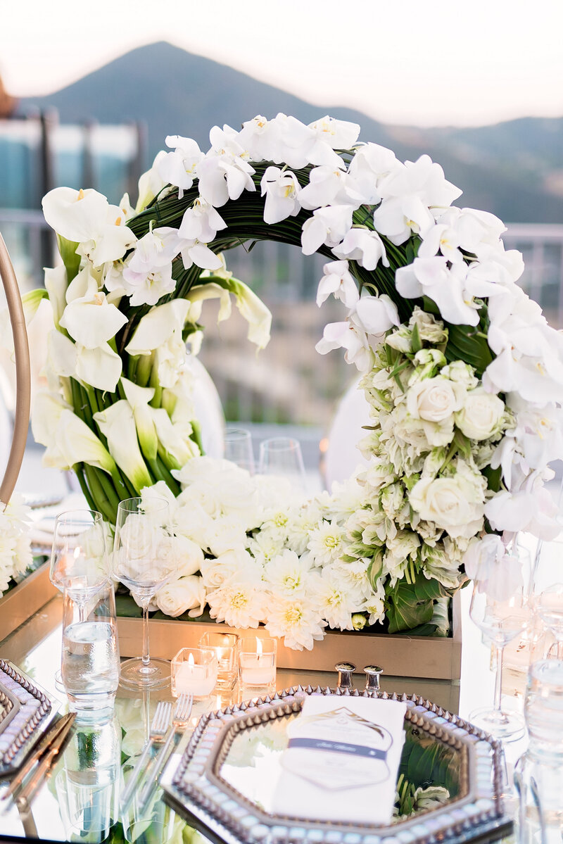 Silver wedding place-setting on a mirrored table with a white floral centerpiece curved into a circular hoop shape.