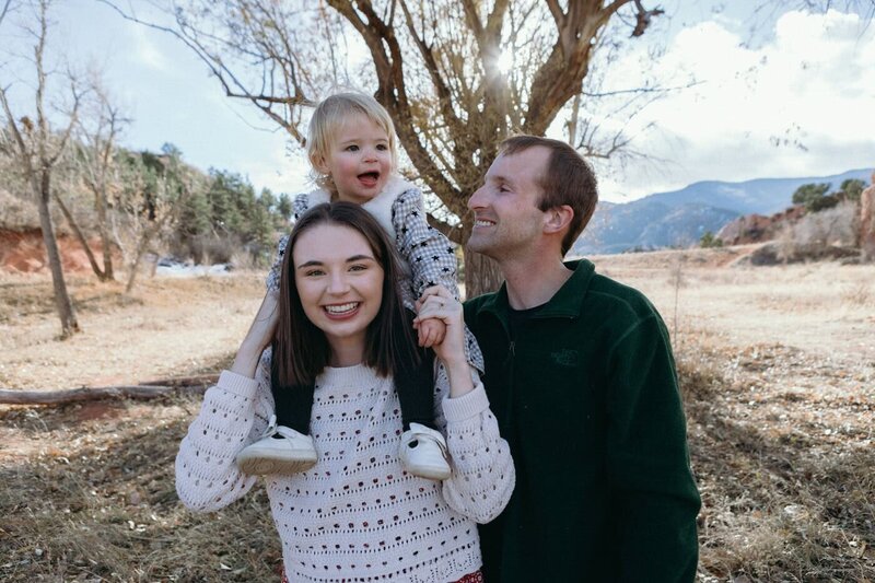 A toddler girl rides on her mom's shoulders while dad gives her a big smile.