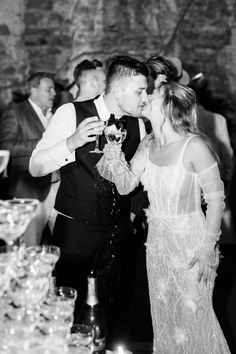 Black and white photo of a bride and groom clinking glasses and leaning in for a kiss during a champagne tower celebration, with the groom spilling his drink.