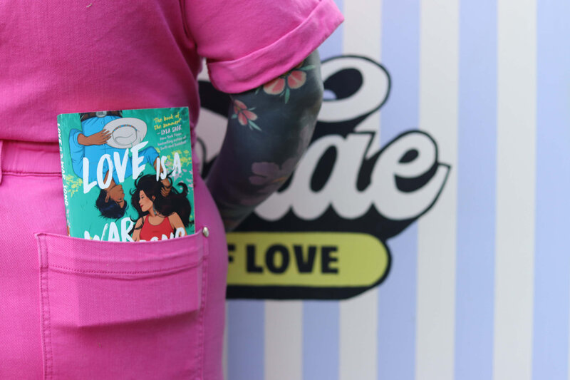 Mid-section of a womans body wearing a pink jumpsuit. There is a romance book in the pocket of the jumpsuit.