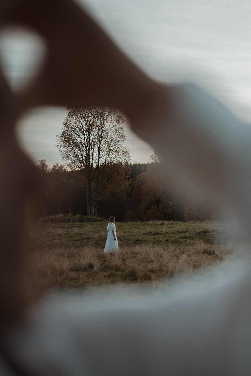 Brudpar står på en äng, bruden står längre bort i det höga gräset och blickar ut