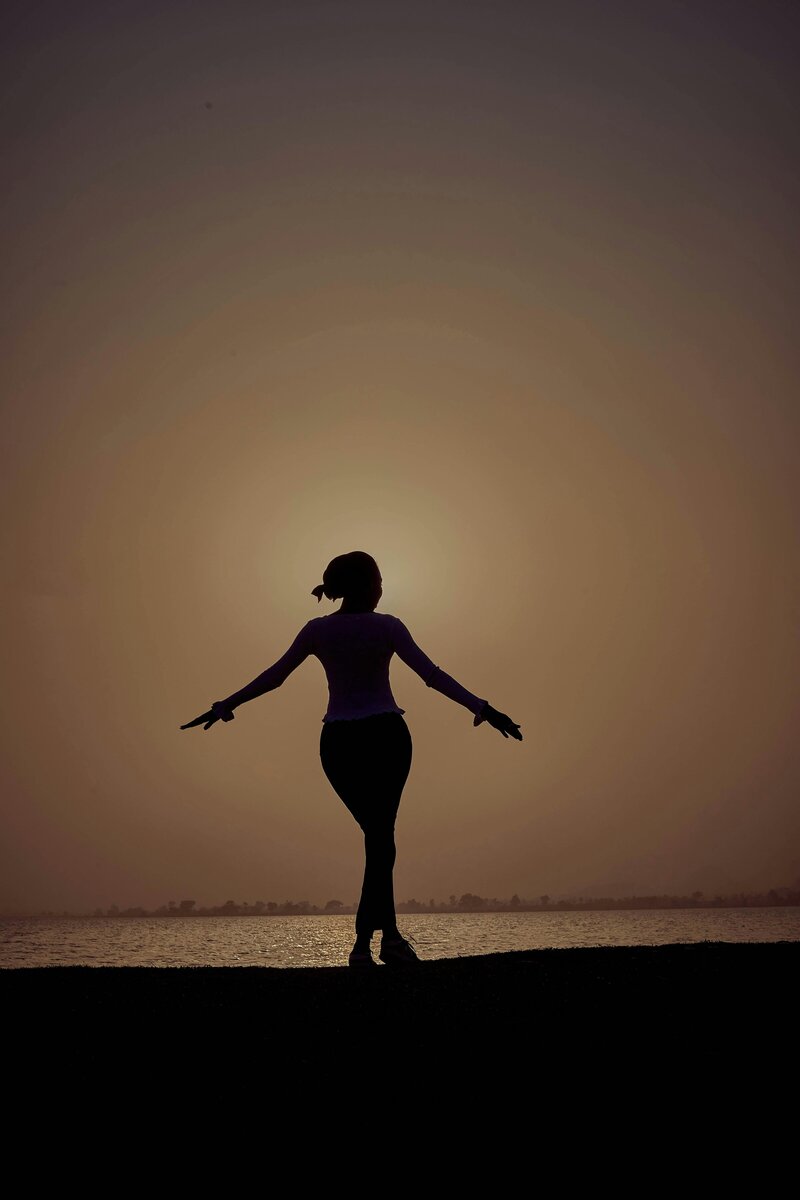 Silhouette of a woman balancing with arms outstretched as she walks on the shoreline with a warm sunset behind her.