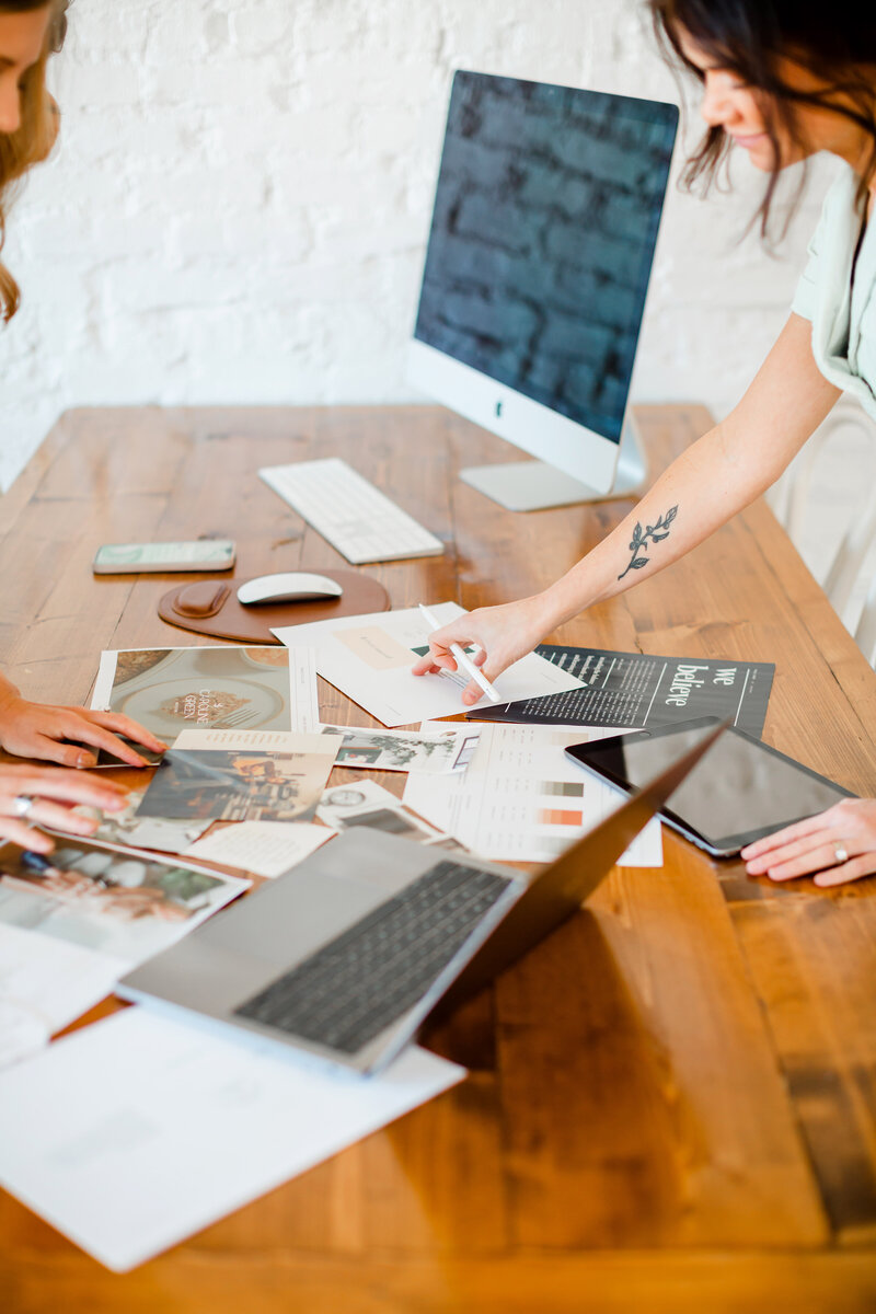 Two women business owners shuffle papers on a wooden desk with a Mac monitor, mac laptop, and iPad | Sara Coffin Photo in NC