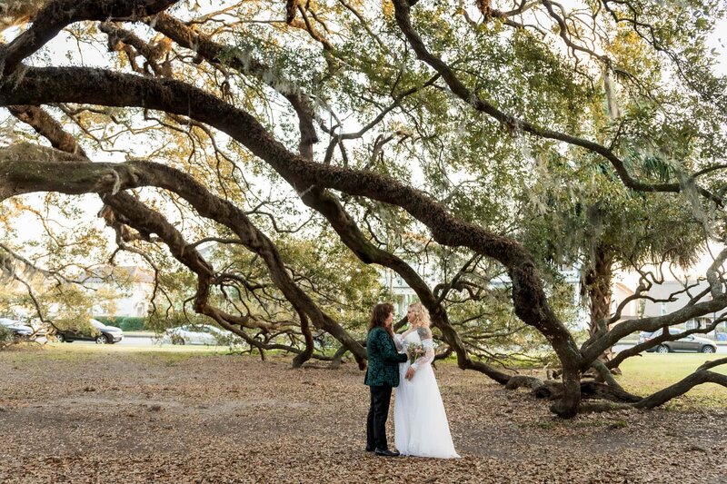 Beneath a canopy of low, winding tree branches draped in Spanish moss, a couple shares a quiet, intimate moment. The bride wears a flowing off-the-shoulder gown, and her partner touches her face lovingly. The setting, part of a destination wedding in the deep South, feels almost magical—surrounded by sprawling oaks, fallen leaves, and golden evening light.