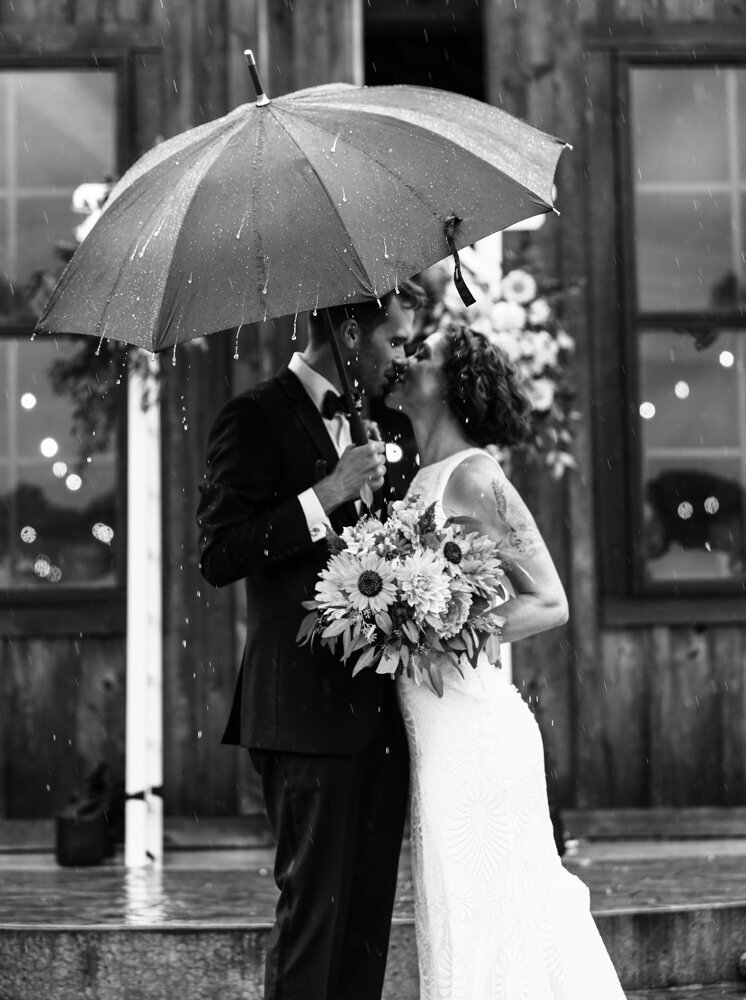 Bride and groom stand  under an umbrella in a rainstorm at the CHQ Barn in Chautauqua NY