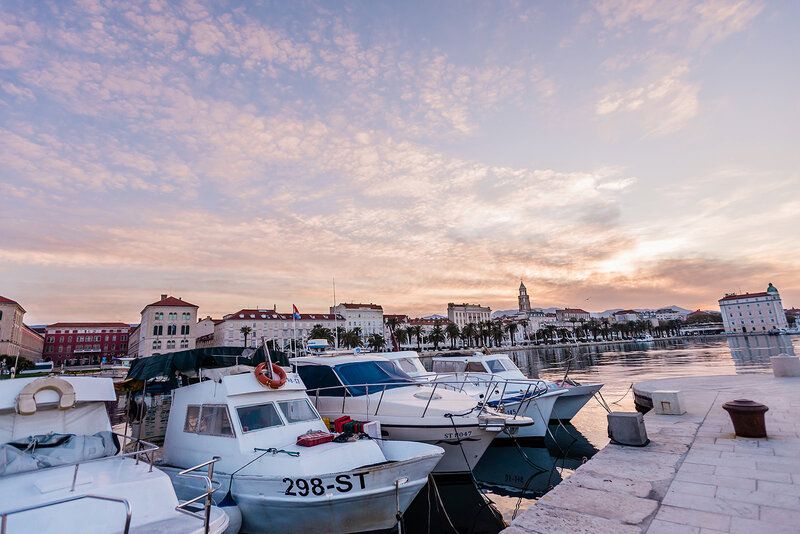 a line up of beautiful white boats line up in harbor next to a marble sidewalk.  The photographer captures the warm orange sunrise with dynamic clouds during a luxury elopement wedding  in Split Croatia.