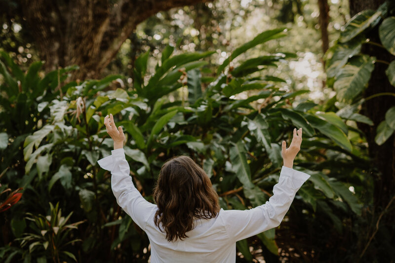 Intuitive healer, Brandi of The Intuitive Momma, raises her arms in a white button down with greenery in the background.