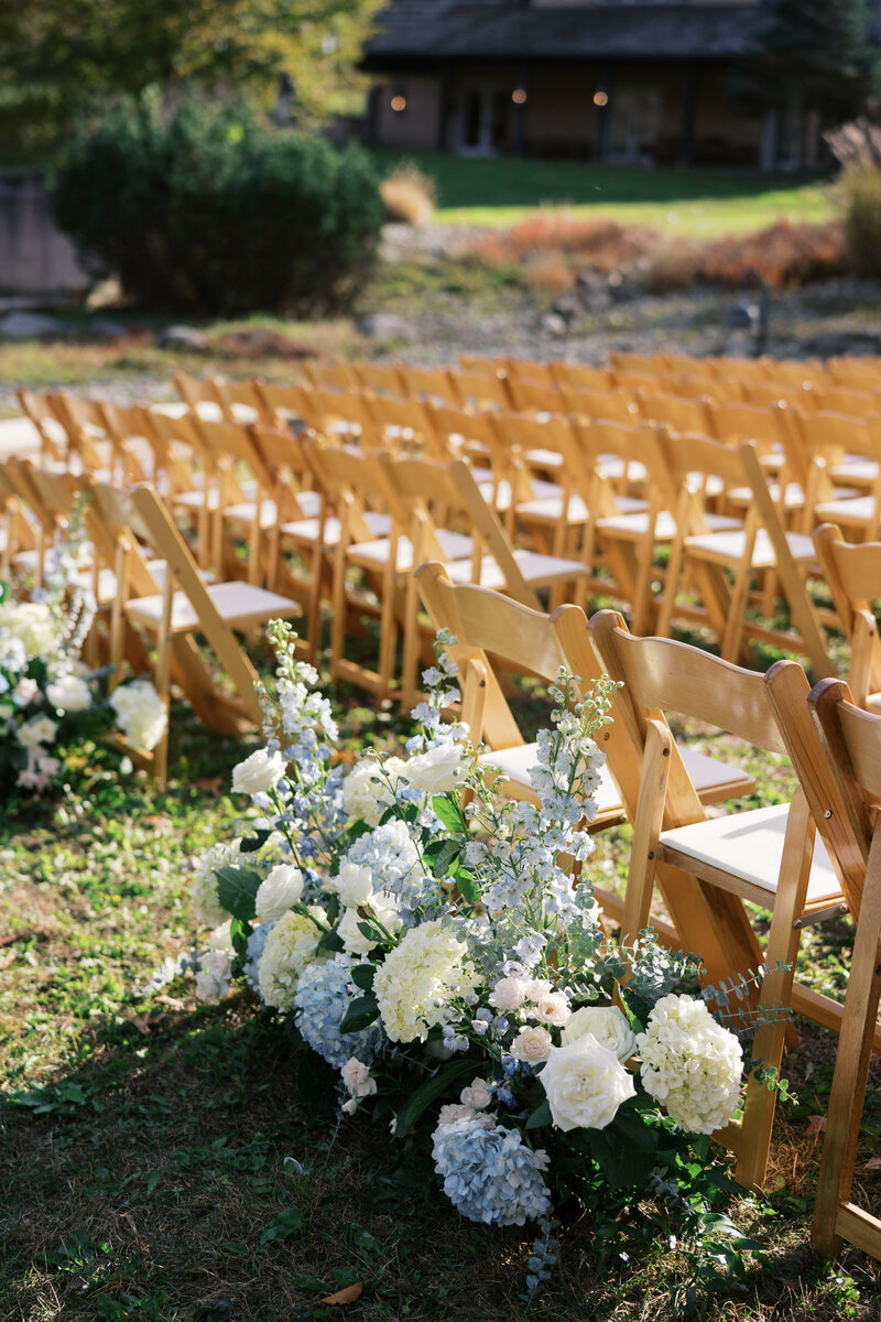 Rows of wooden ceremony chairs adorned with blue and white floral arrangements in an outdoor setting.