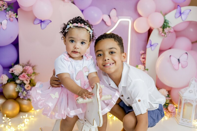 Two children posing for a photo at a one-year-old birthday party in front of a balloon wall and neon number 1.