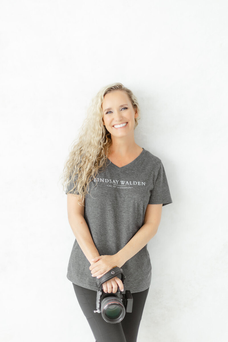 Lindsay Walden, Dallas fine art photographer, smiling and holding her camera while wearing a gray studio t-shirt against a bright white background.