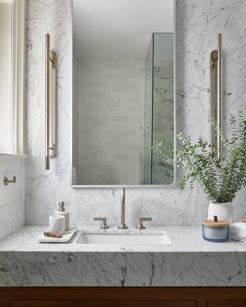 A luxury bathroom vanity with a marble countertop and soap bottles, brushed nickel fixtures, a large mirror, and potted plant next to a bright window