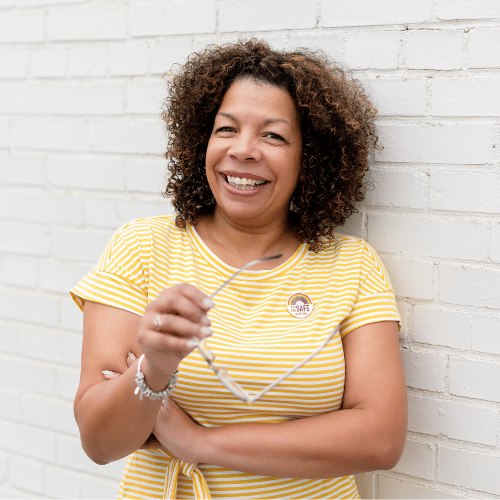 Photo of Michelle McKown-Campbell smiling in a yellow and white striped dress with a "Safe" LGBTQ rainbow button and holding her eye glasses