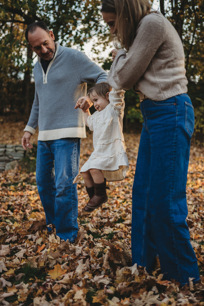 A joyful family plays in fallen autumn leaves as parents swing their young child between them during a fall family photography session in Connecticut. Captured by Nicole Mele Photography, showcasing warm and candid moments.