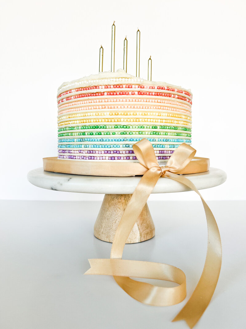 Round cake with rows of rainbow colored sprinkles, topped with candles.