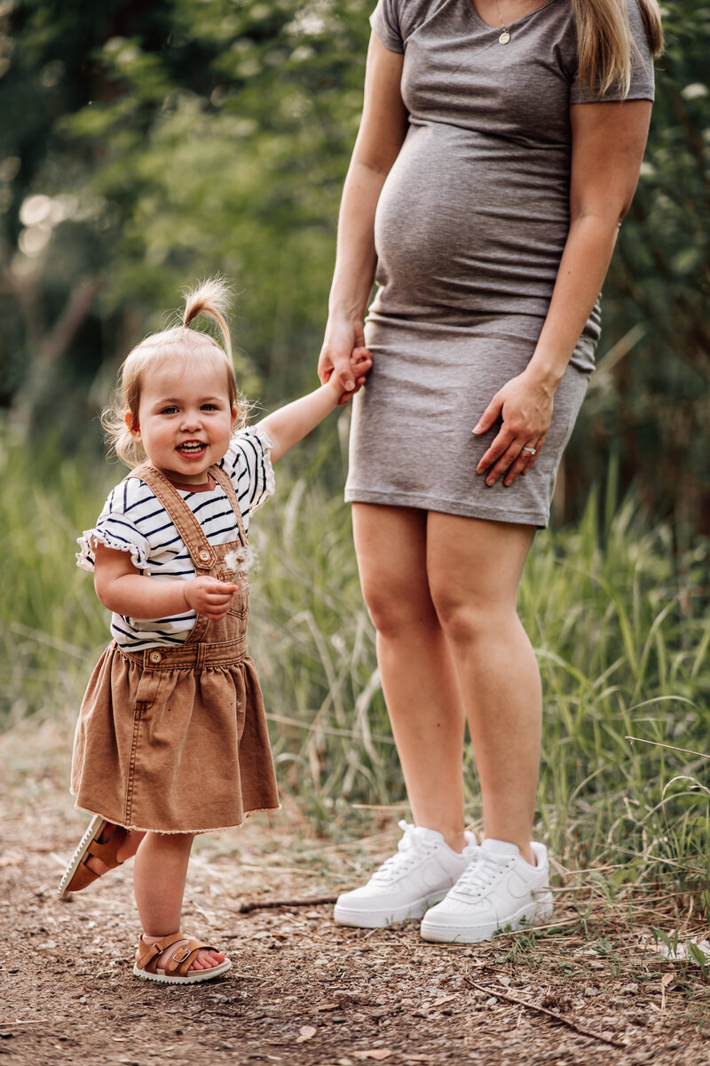 A pregnant woman in a gray dress holds hands with a smiling toddler who is holding a dandelion. The toddler is wearing a striped shirt and a brown skirt overall, standing on a dirt path surrounded by greenery.