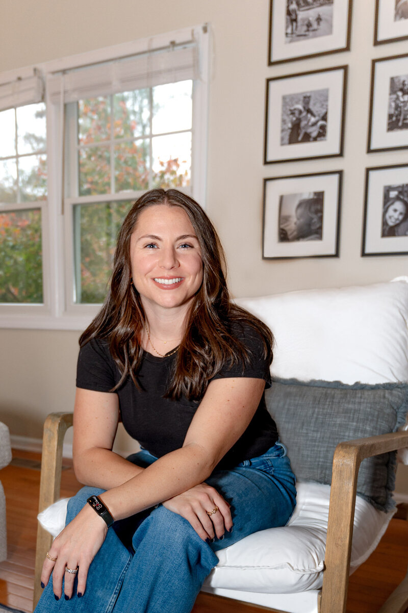 Female consultant posing for a headshot, leaning forward while sitting in a white chair, smiling at the camera in her home in Weston, CT.