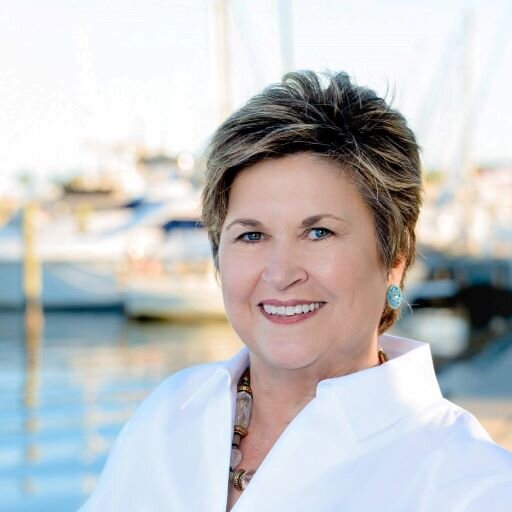 Carolyn Quintin, midlife career coach, poses in front of a body of water with sailboats in the background. She is wearing a