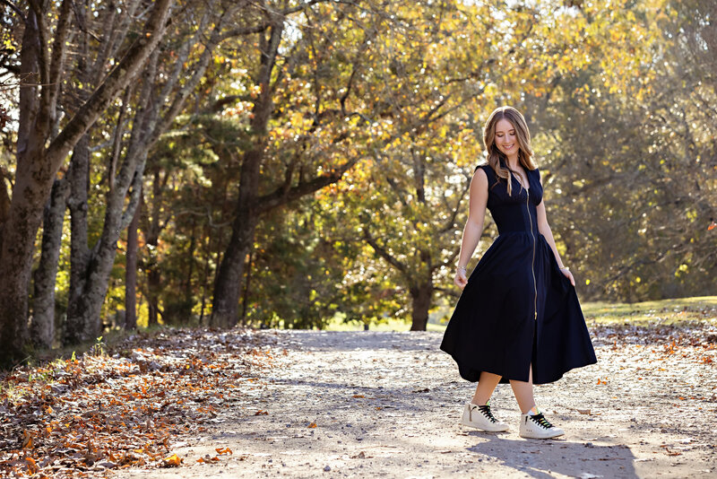 High school senior in black dress walking dreamily along gravel path lined with yellow trees