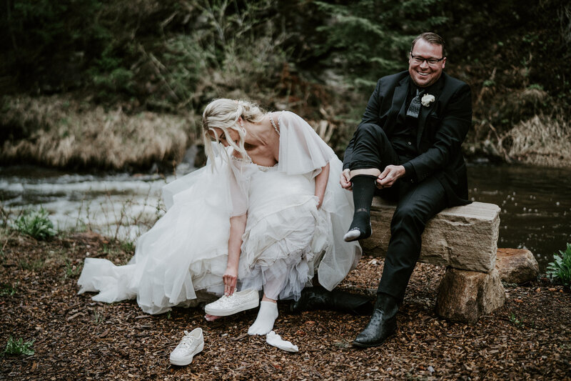 Bride and groom laughing while changing into sneakers by a creek at their Breckenridge elopement.