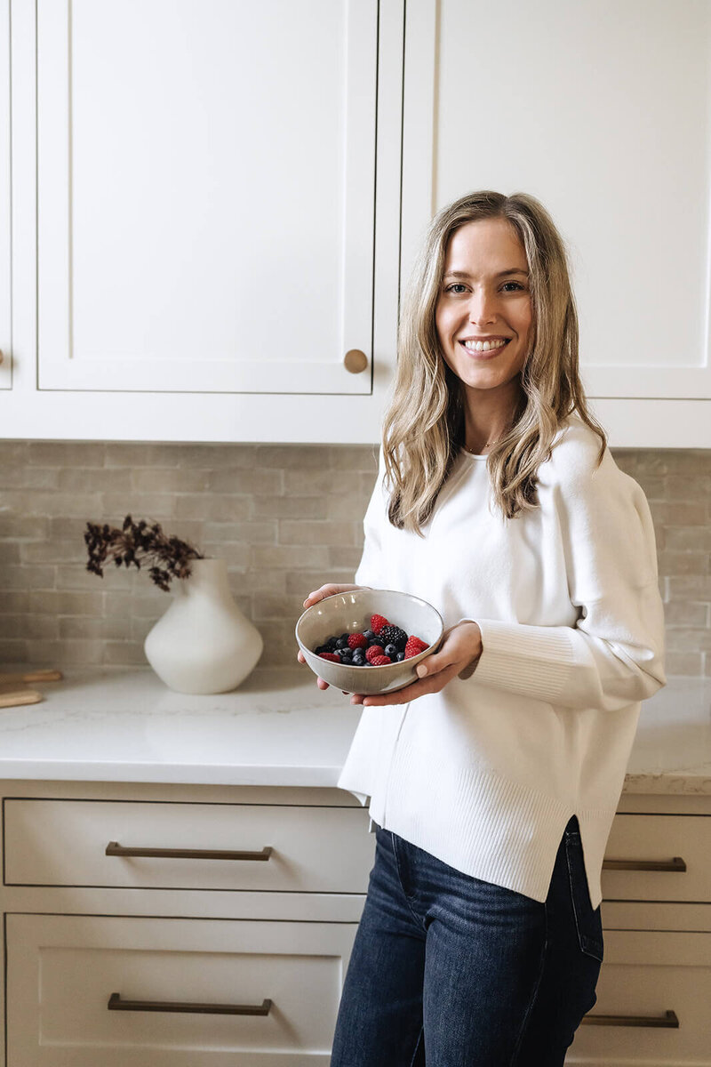 Smiling wellness coach for moms holding a bowl of fresh berries in a modern kitchen, promoting simple healthy eating for early motherhood