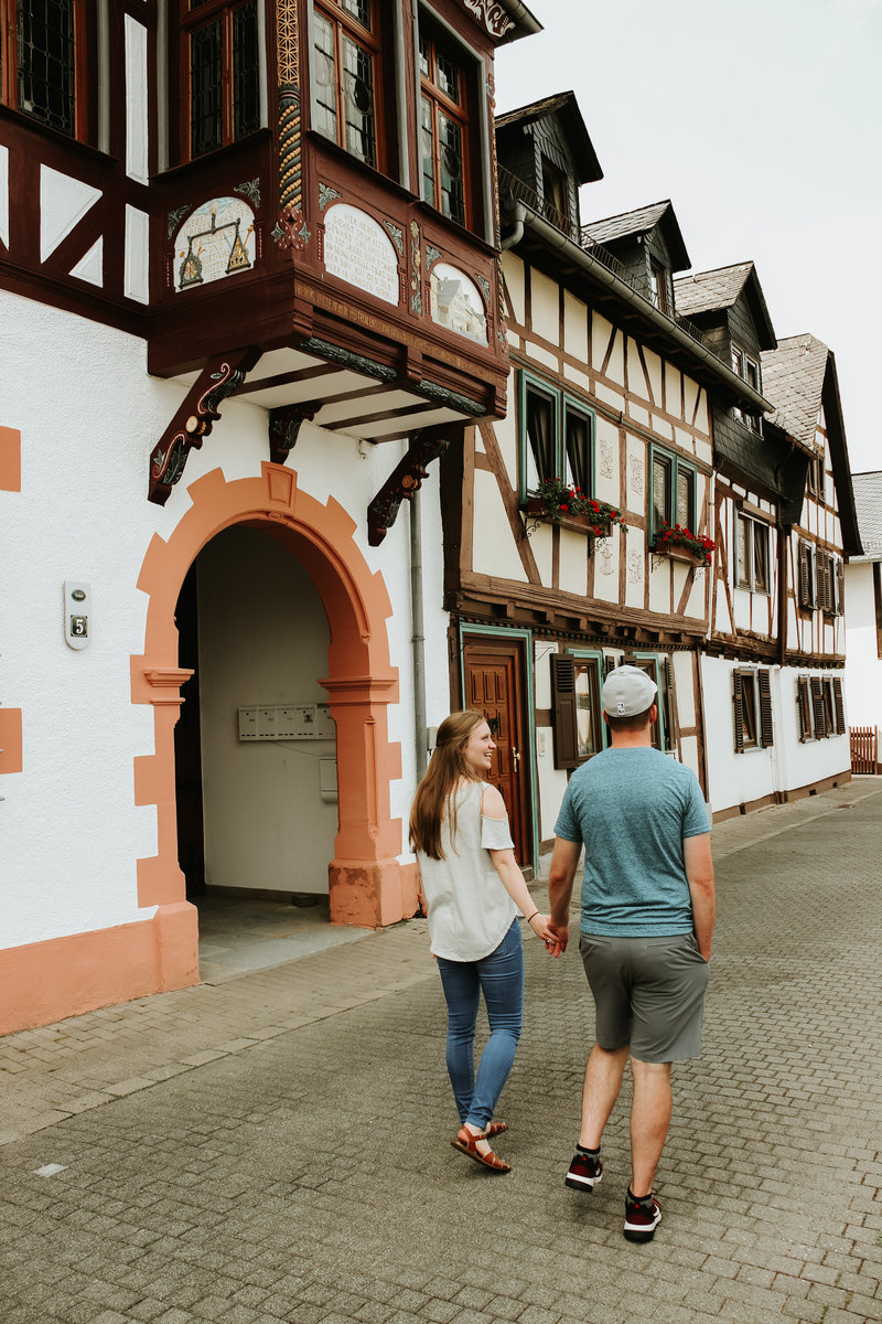 A young couple walk holding hands next to a timber, German building