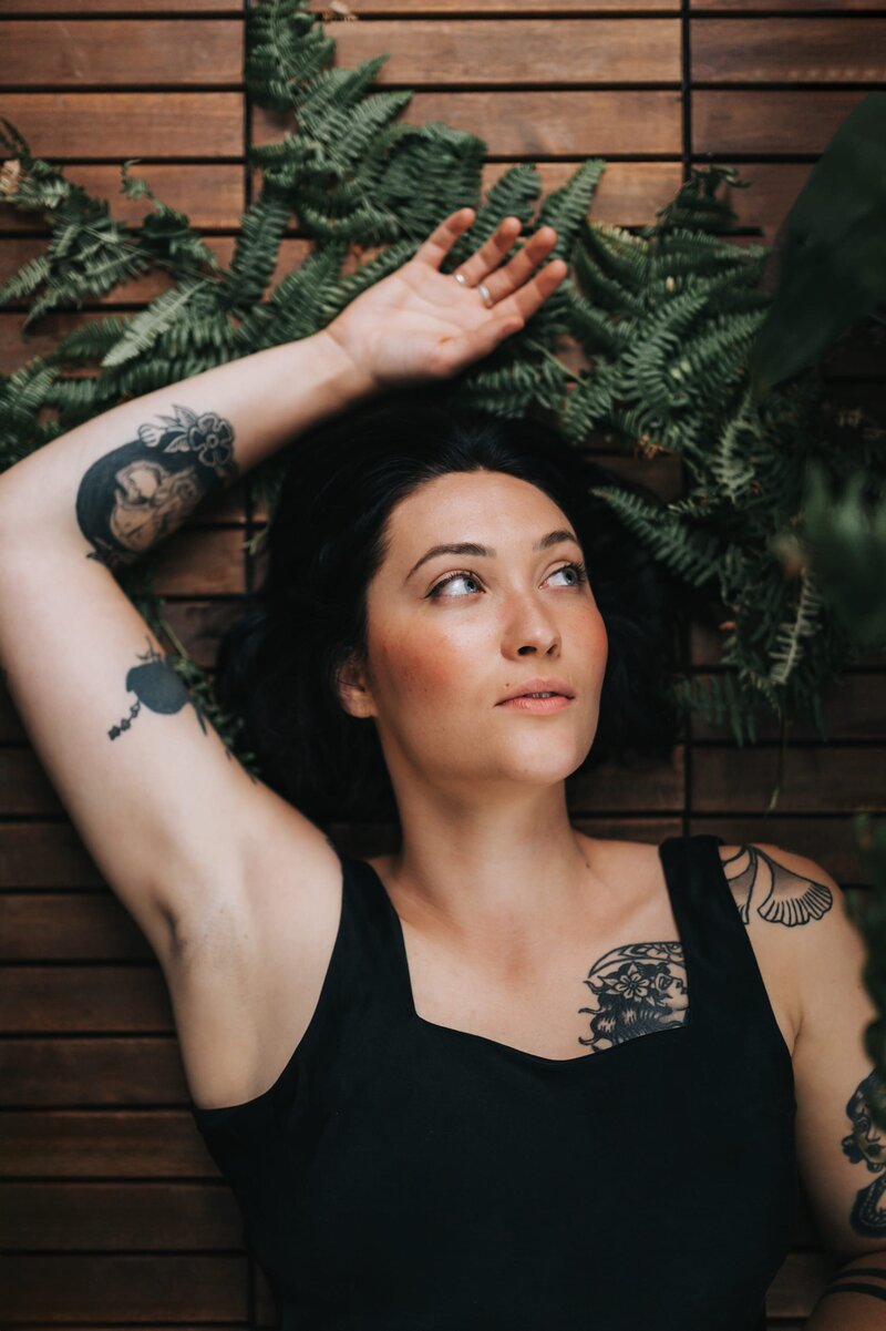 Woman with tattoos lays atop several ferns with her arm stretched above her.