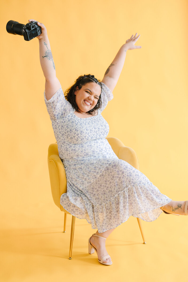 A person sitting in a yellow chair with both hands in the air and a camera in one of them