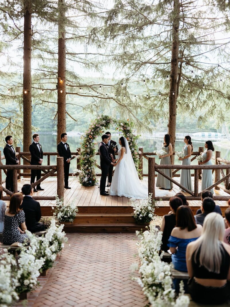 Groom and Bride stand in the center of their romantic wedding ceremony at Cedar Lakes Estate venue in Hudson Valley. Aisle lined with white flowers, guests watch as bridal party stands on wood deck under twinkle lights. Elegant floral arbor in background with pine trees and lake.