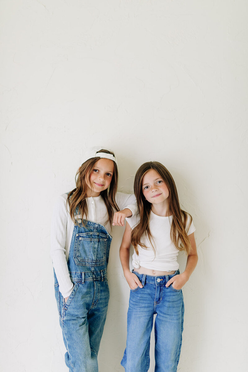 Two sisters ages seven and ten, posing on a white wall wearing denim