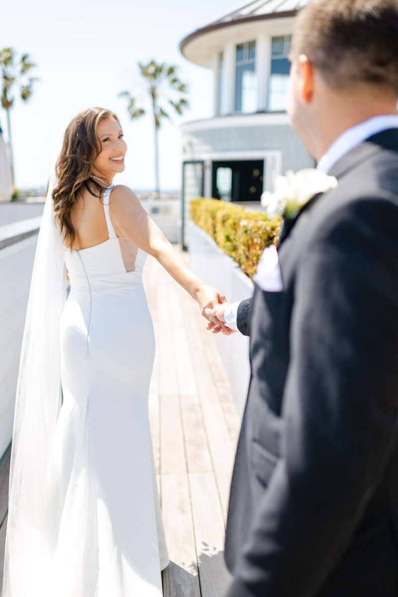 Bride and groom at Lido House in Newport Beach, holding hands on the rooftop deck with ocean views. The bride smiles over her shoulder as the groom follows, creating a romantic and timeless wedding moment