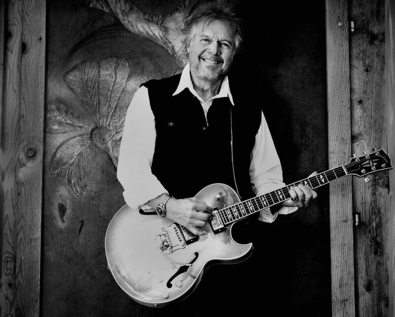 Randy Bachman black and white portrait smiling while holding guitar against wood wall with flower engraveing