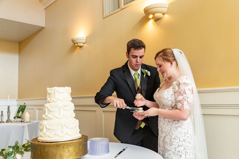 A bride and groom, dressed in wedding attire, smile as they cut and serve each other a piece of a three-tiered white wedding cake during their reception in a softly lit room.