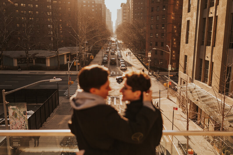 A couple shares an intimate moment, slightly blurred, with a backdrop of a sunlit New York City street lined with tall buildings.