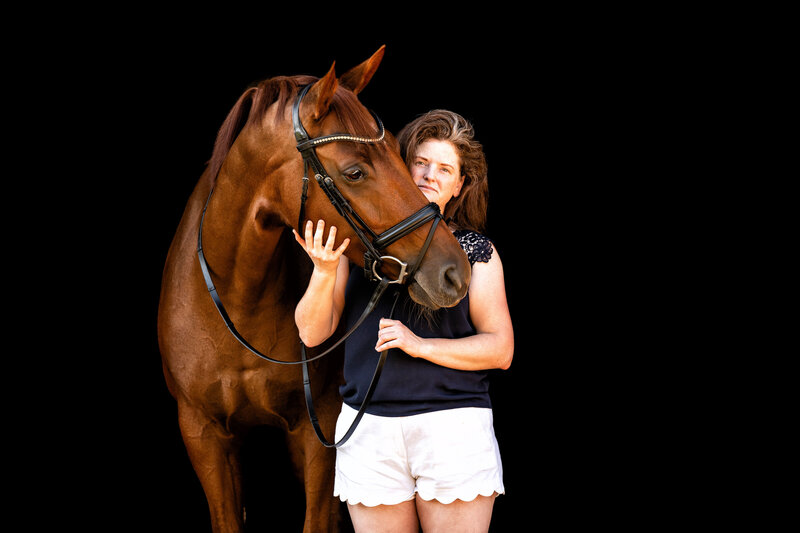 Chestnut horse puts head over woman's shoulder on black background.