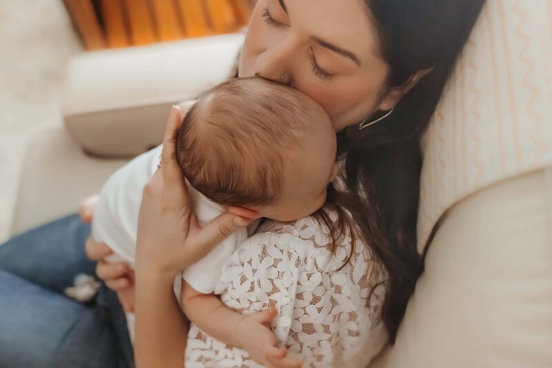 Woman sitting on a couch, gently holding and kissing the head of a baby resting on her chest.
