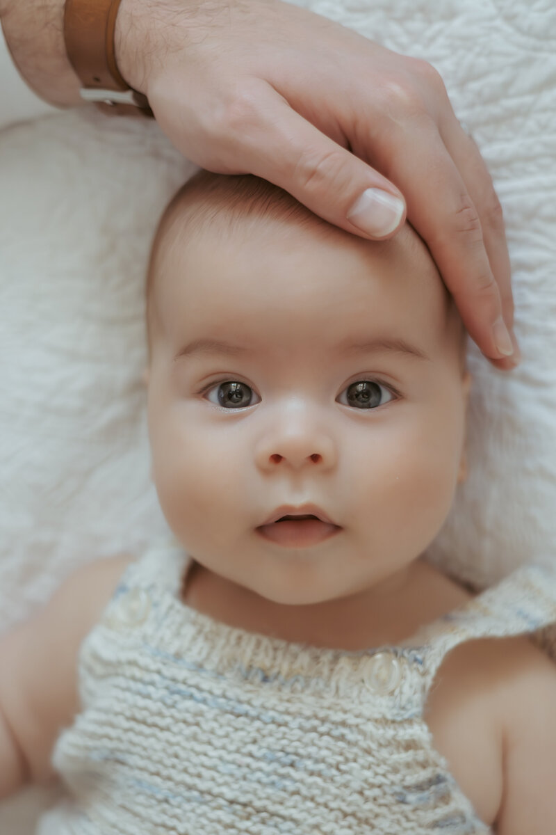 A four month old baby in a knitted romper is lying on bed with his dad hands on his head and tummy.